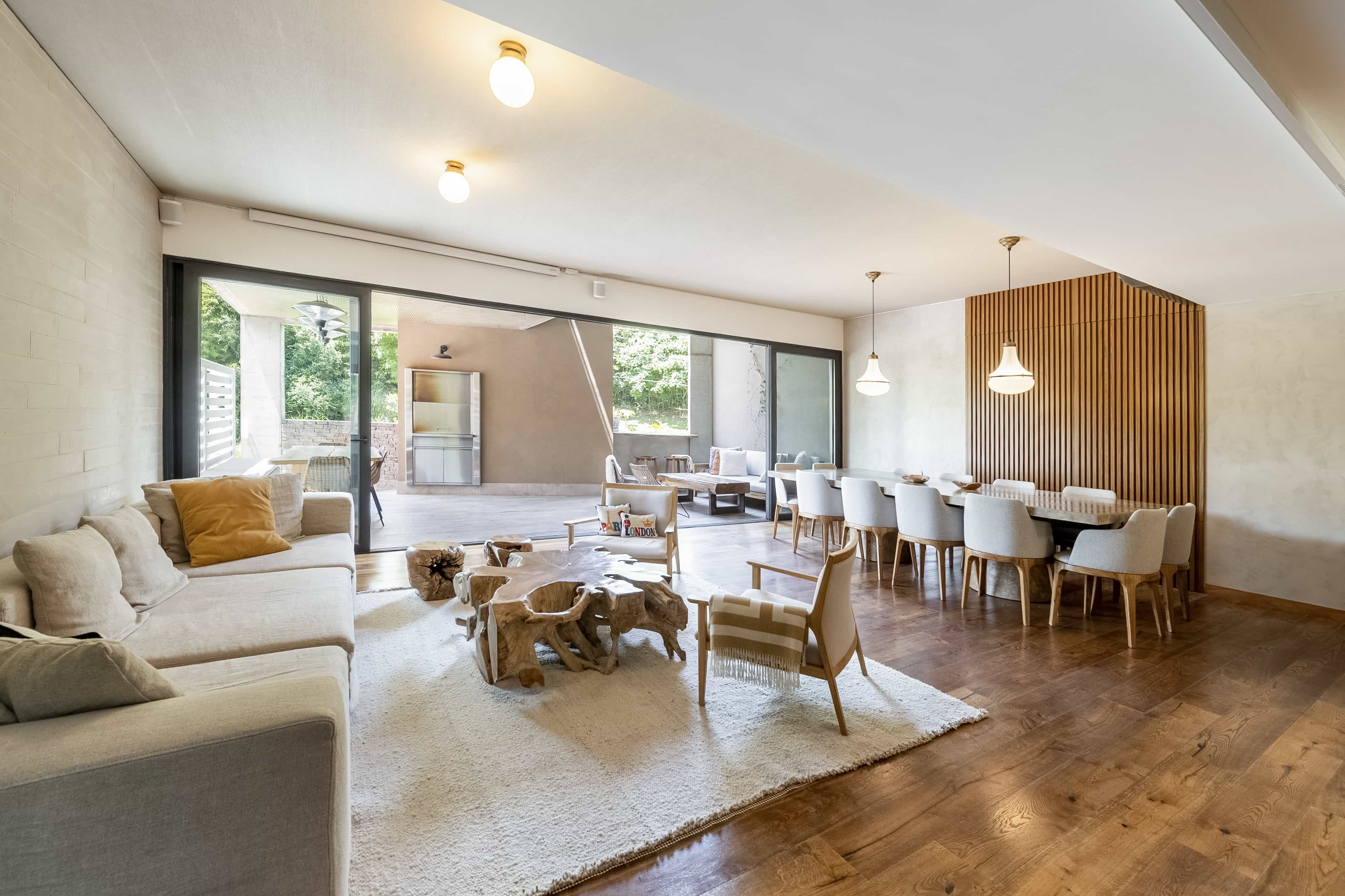 Casa con un gran living-comedor. Sobre la izquierda se encuentra un sofá con almohadones grises y naranja. En el centro una mesa realizada con un tronco de una arbol. Sobre la derecha una gran mesa con sillas blancas.